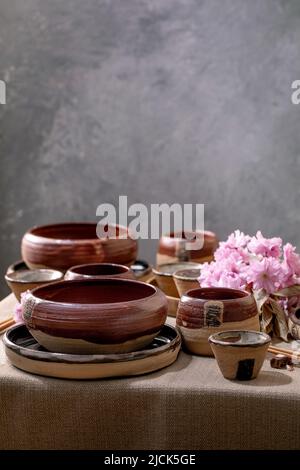 Japanese Asian style table setting with empty craft ceramic tableware, brown rough bowls and cups on linen tablecloth, decorated by pink spring flower Stock Photo