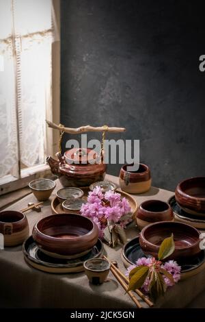 Japanese Asian style table setting with empty craft ceramic tableware, brown rough bowls, craft teapot and cups on linen tablecloth, decorated by pink Stock Photo