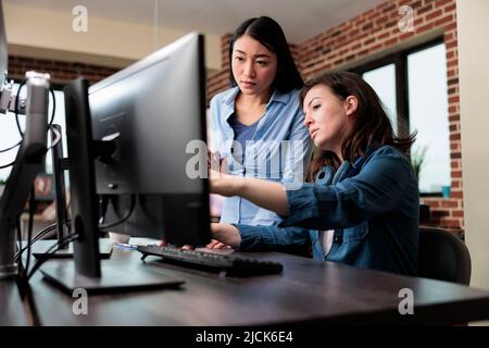 Professional office workers pointing out wrong blueprint of project while using work computer. Creative designers developing digital arts on PC while discussing about 3D technology. Stock Photo