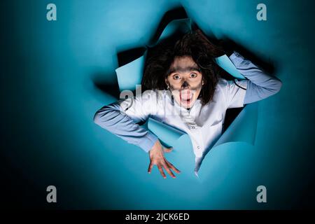 Foolish insane scientist with wacky look and dirty face after laboratory explosion acting crazy on blue background. Lunatic silly chemist with messy hairstyle, looking funny while screaming at camera. Stock Photo