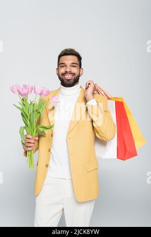 positive african american man in yellow blazer and polo neck holding pink tulips and shopping bags isolated on grey Stock Photo