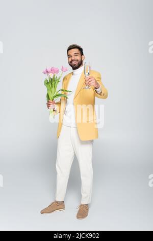 full length of happy african american man in yellow blazer holding pink tulips and glass of champagne isolated on grey Stock Photo