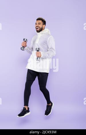 full length of excited african american man in sportswear levitating and holding dumbbells on purple Stock Photo
