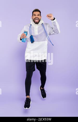full length of excited african american man in sportswear levitating and holding sports bottle on purple Stock Photo