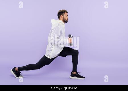 full length of bearded african american man doing lunges with dumbbells on purple Stock Photo