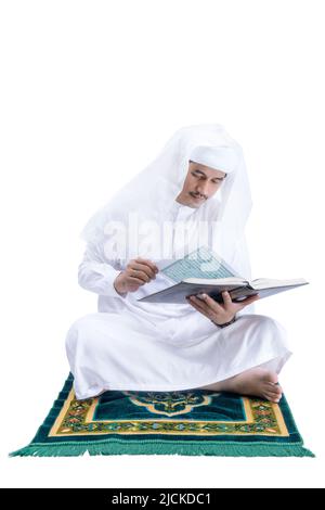 Arab Muslim man sitting and reading the Quran on the prayer rug isolated over white background Stock Photo