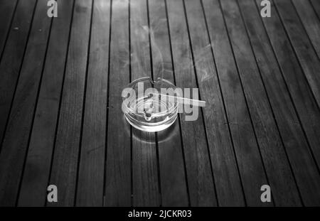 Burning cigarette in a glass ashtray on a wooden table. Black and white. Stock Photo