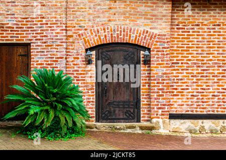 A closed old wooden massive door with a knock on a doorknob in the form of a twisted ring, vintage lanterns hang on both sides of a red brick wall. Stock Photo