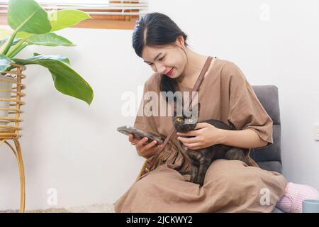 Pet lover concept, Young Asian woman using smartphone and playing with cat while relaxation at home. Stock Photo