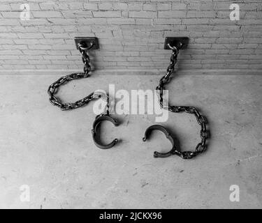 A set of empty old iron shackles draped on the floor connected to an isolated white brick wall  - 3D render Stock Photo