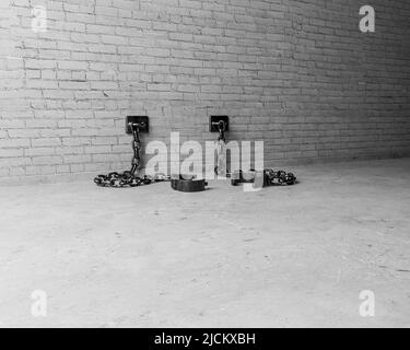 A set of empty old iron shackles draped on the floor connected to an isolated white brick wall  - 3D render Stock Photo