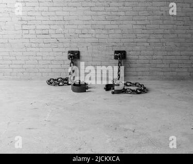 A set of empty old iron shackles draped on the floor connected to an isolated white brick wall  - 3D render Stock Photo