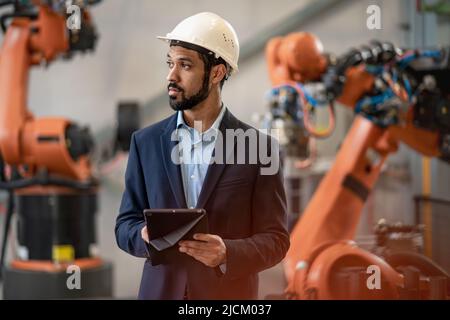 Automation engineer uses tablet for programming robotic arm in factory. Stock Photo