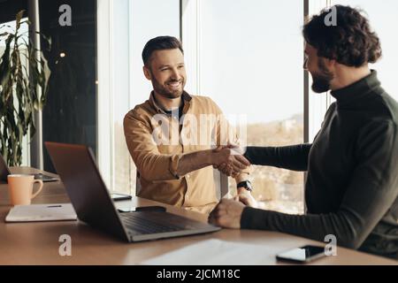 Successful businessmen shaking hands in agreement, sitting at desk with laptop at company office, free space Stock Photo