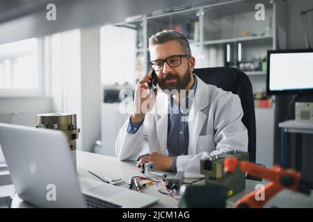 Robotics engineer calling on cellphone and working on desing of modern robotic arm in laboratory office. Stock Photo