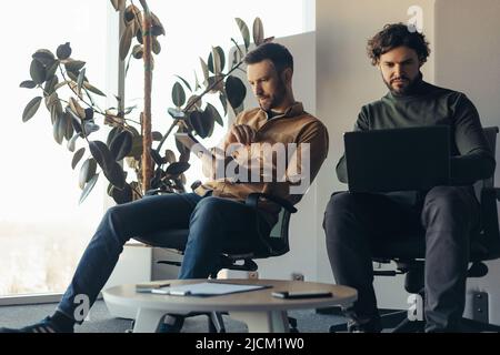 Serious male colleagues working together, using laptop computer and digital tablet at contemporary company office Stock Photo