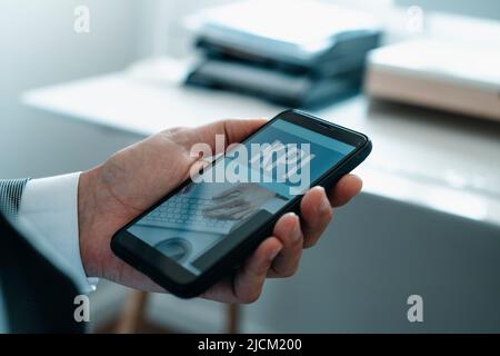 closeup a man at the office, wearing a formal white shirt and a gray suit jacket, holding his smartphone, that reads the text KPI, for key performance Stock Photo
