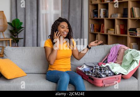 Excited black woman sitting near open suitcase with clothes, speaking on cellphone, booking travel tour from home Stock Photo