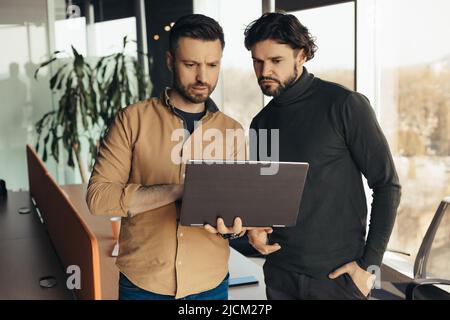 Two focused male employees developing business strategies, cooperating with each other, using laptop pc at modern office Stock Photo