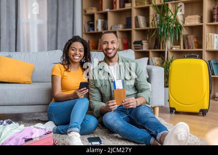 Happy black spouses booking tour online, using cellphone, man holding passport with tickets, smiling together to camera Stock Photo