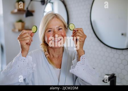 Beautiful senior woman in bathrobe applying cucumber face mask in bathroom, skin care concept. Stock Photo
