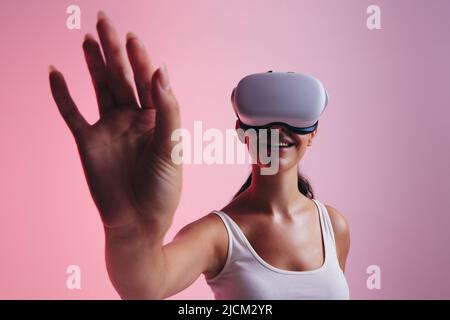 Cheerful young woman using her hand to interact with virtual space while wearing virtual reality goggles. Happy young woman experiencing a 3D simulati Stock Photo