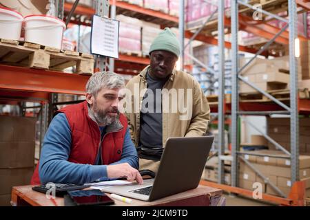 Mature bearded manager using laptop to check online transportation together with his colleague standing behind him at warehouse Stock Photo