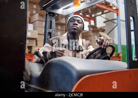 Serious African worker in uniform driving forklift at warehouse to load cargo Stock Photo