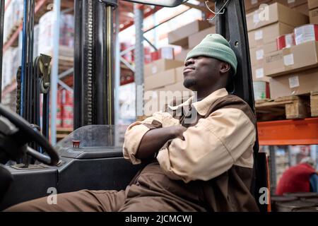 African young driver of forklift sitting on seat and relaxing after work at warehouse Stock Photo