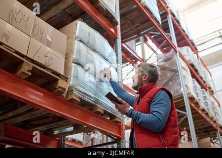 Mature worker using tablet pc and noting packages on shelves at warehouse before shipping Stock Photo