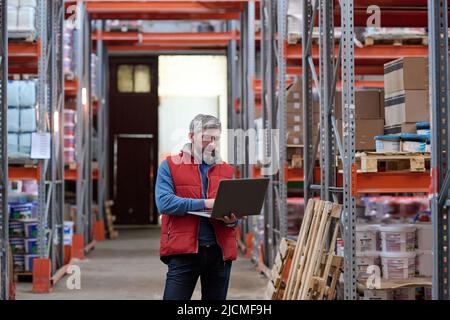 Mature bearded distributor using laptop while walking along the shelves with parcels at warehouse Stock Photo