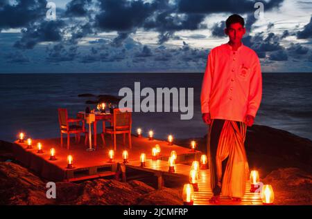 A waiter lights candles and sets a table for two for a romantic dinner on the beach. Aturuwella, Bentota, Sri Lanka. Stock Photo