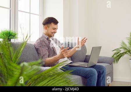 Happy young man sitting on couch at home and having video call on laptop computer Stock Photo
