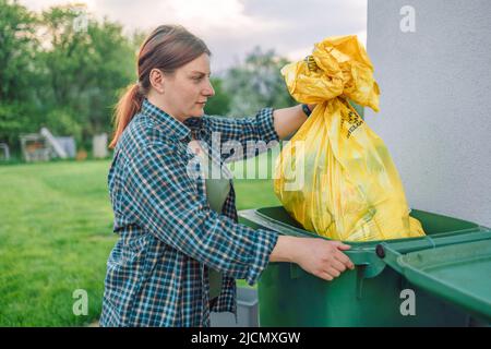 European 30s woman throwing garbage into the recycling bin in the backyard near the house. Separating recyclable garbage plastic  Stock Photo