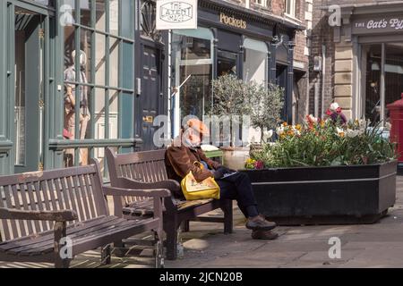 A middle aged man wearing a cap is writing in a book while sitting on a bench in the street. Stock Photo