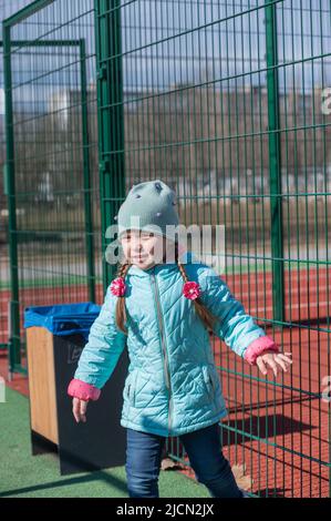 Portrait of a child having fun on a sports field. A five-year-old girl with long hair. Hair braided into pigtails. The child is dressed in warm clothe Stock Photo