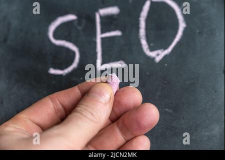 Adult male holding a piece of colored chalk in his hand. Handwritten word SEO on black chalkboard. Stock Photo