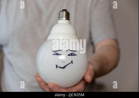 A middle-aged man is holding an electric lamp with his face painted on it. A happy smiling face is drawn in black marker. A large round electric lamp Stock Photo