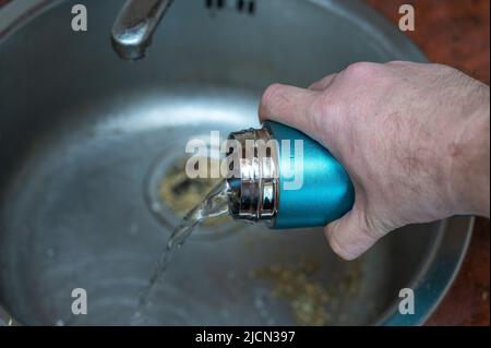 A man pours herbal tea from a vacuum flask into the sink. An adult man holds a cylindrical turquoise thermos in his hand. Container for hot and cold d Stock Photo