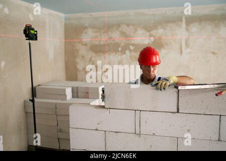 Bricklayer using spirit bubble and laser level to precise check concrete blocks on wall. Contractor uses tools for brickwork. Worker constructs a wall Stock Photo