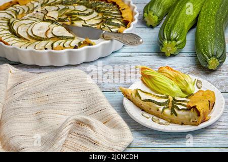 Open zucchini tart with herbs and cheese curd filling in a baking dish and sliced on a wooden table surrounded by ingredients Stock Photo