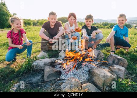 Group of Kids - Boys and girls cheerfully smiling and roasting sausages on long sticks over a campfire flame. Outdoor active time spending or camping Stock Photo