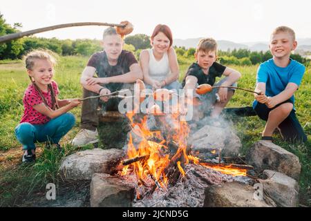 Group of Kids - Boys and girls cheerfully smiling and roasting sausages on sticks over a campfire flame Sausages selective focus. Outdoor active time Stock Photo