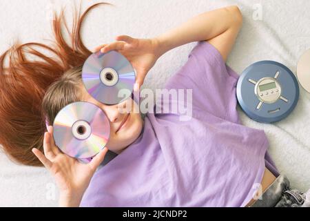 Happy teenage girl in lilac t-shirt lies on bed in pink headphones holds cd disks in her hands Stock Photo