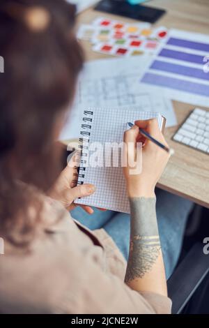 Close up of female interior designer maing notes in her notebook while working on a project with blueprint and color samples, sitting at the desk in Stock Photo