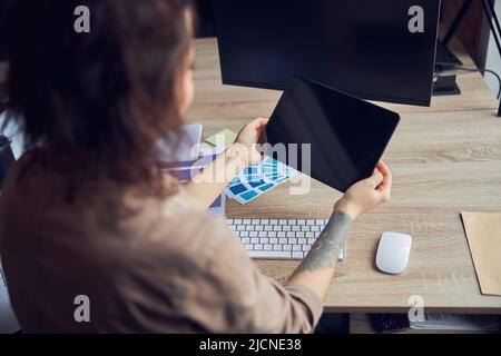 Rear view of female interior designer holding tablet pc with blank screen while working on a project with color samples, sitting at the desk in her Stock Photo