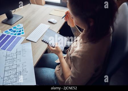 Female interior designer holding pencil, making notes in her notebook while working on a project with blueprint and color samples, sitting at the desk Stock Photo