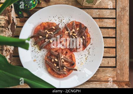 Organic tomato slices covered with anchovies and pine nuts on a teak wood table Stock Photo