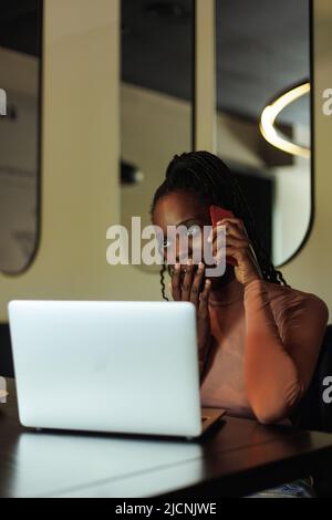 Vertical shocked, surprised multiethnic business woman covering mouth with hand, talk phone. Remote online work in cafe  Stock Photo