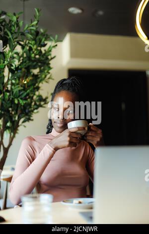 Vertical photo of calm, contemplating African American businesswoman with laptop, drink coffee or tea in internet cafe Stock Photo